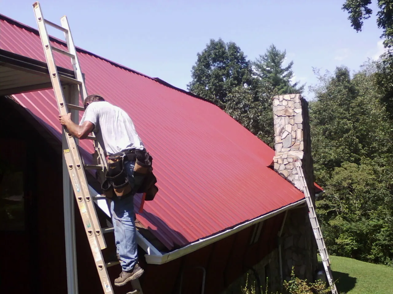 Roofer on a ladder beside a red metal roof and stone chimney — Soffit & Fascia Repair by Roofing Brothers in Seibert